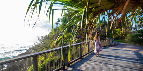 A girl in dress walks along the boardwalk and enjoys the view of the stunning Coolum Beach and surfers catching big waves, Pacific Ocean. Stunning panorama of Sunshine Coast, Queensland, Australia