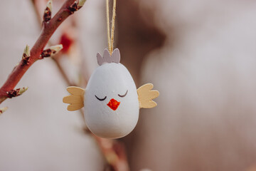 Easter egg in a form of a cute chicken hanging on a branch of a flowering tree in a spring garden in sunny day. Easter wallpaper for desktop. Greeting card. Decor for a springtime religious holiday.