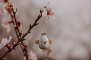 Easter egg in a form of a cute chicken hanging on a branch of a flowering tree in a spring garden...