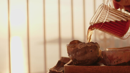 Pouring chinese pu-erh tea from glass mug into beautiful cup for tea and drinks. Morning or evening tea ceremony at sunset or dawn in street with sunbeams.