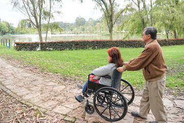 Senior hispanic couple strolling in a park, he pushes her wheelchair along a pathway.