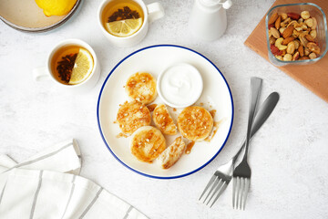 Plate with tasty cottage cheese pancakes, sour cream and cups of tea on light table