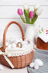 Fototapeta na wymiar Wicker basket with festively decorated eggs, beautiful tulips and traditional Easter cake on white wooden table