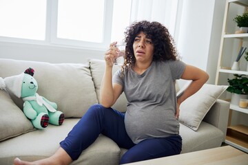 A pregnant woman sits on the couch with lower back pain and headache, strain on during pregnancy, drinks water and looks at the laptop. Lifestyle preparation for childbirth, last month of pregnancy