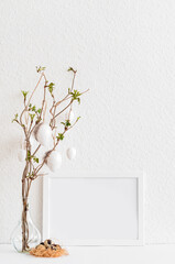 Vase with tree branches, Easter eggs and blank picture frame on table near light wall