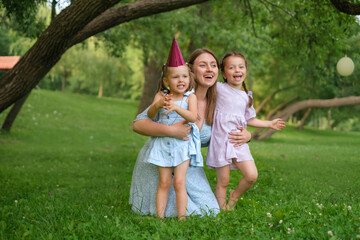 Fototapeta premium Mom hugs her daughters and laughs merrily sitting in the park on the lawn. A birthday girl with a festive hat on her head. Mothers Day.