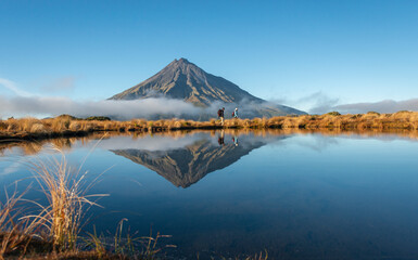 Two people hiking Pouakai circuit. Mt Taranaki reflected in the Pouakai tarn. New Zealand.