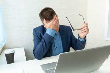 Tired or exhausted man closing his face with hands sitting in front of computer in office....