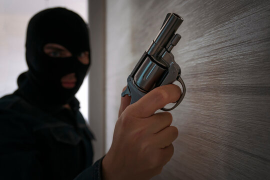 Terrorist hooded in balaclava, holding gun in hand. Man aiming with a pistol, dark building indoor background, Killer, armed robbery concept.