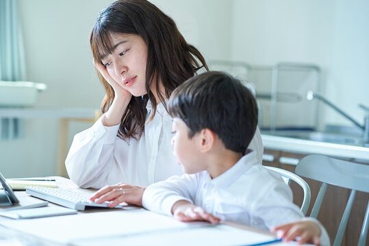 部屋で遊ぶ子供と、隣で仕事をして疲れた表情の母親