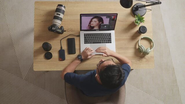 Top View Of A Male Editor Walking Into The Workspace Sitting Down And Using A Laptop Next To The Camera Editing Photo Of A Female At Home

