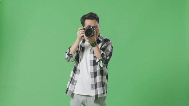 Asian Photographer Using A Camera Taking Pictures And Smiling Touching His Chest While Standing On Green Screen Background In The Studio
