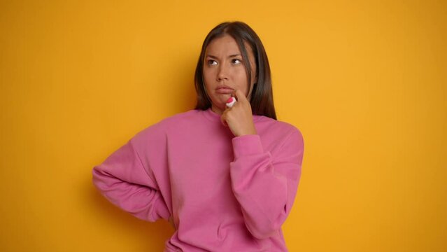 Pretty young brunette girl stands against yellow background, she thinks about somethig and looks up thoughtfully, copy space, high quality video