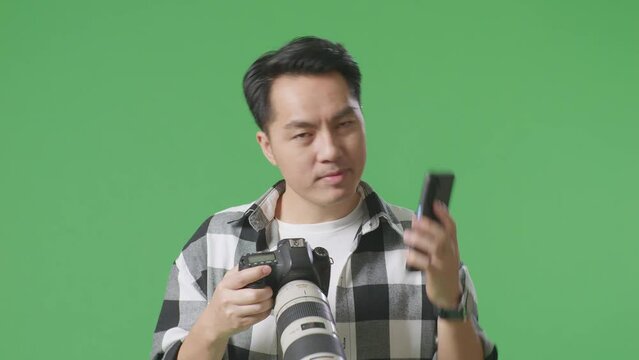 Close Up Of Asian Photographer Holding A Camera In His Hands And Talking On Smartphone While Standing On Green Screen Background In The Studio
