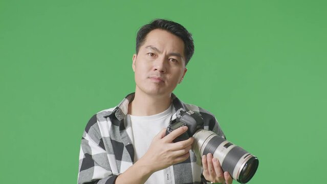 Close Up Of Serious Asian Photographer Holding A Camera In His Hands And Disapproving With No Index Finger Sign While Standing On Green Screen Background In The Studio
