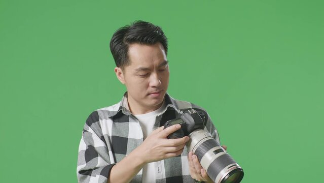 Close Up Of Asian Photographer Looking At The Pictures In The Camera Then Waving His Hand While Standing On Green Screen Background In The Studio

