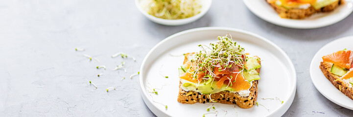 Flat lay top view of healthy sandwich with smoked salmon, cucumber, cream cheese and fresh microgreens alfalfa sprouts on white plate on gray concrete background. Healthy lifestyle. Growing sprouts