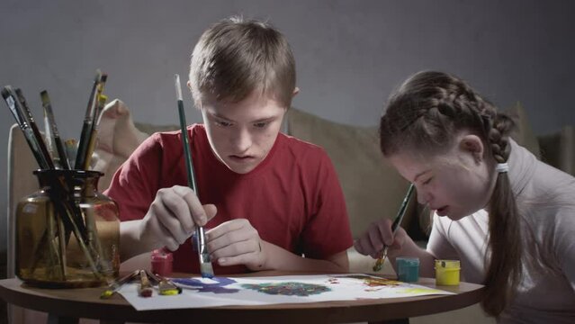 Two children with Down syndrome. A boy and a girl draw with paints and a brush at home. Communication of kids with disabilities. A person with special needs. Chromosomal genetic disorder in a child.