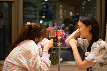 Pretty Asia university female students chat, driink tea or wine near window with blurred bokeh colorful City light background.