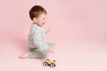 Happy toddler baby is playing with a toy car against a pink background. Child boy rolls a wooden toy car. Kid age one year eight months, full height