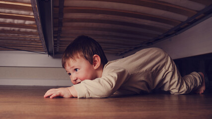 Toddler baby crawls and hides under the bed. Child climbed under the sofa and sits on the floor....