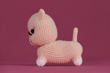 Cute little crocheted cat, handmade art. Amigurumi kitten doll on pink background. A soft DIY toy made of natural cotton and wool. Side view.