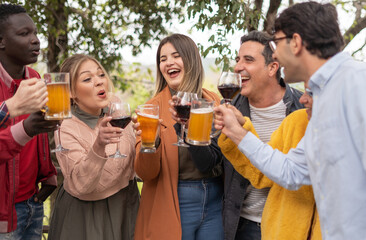 Group of multicultural friends having lunch outdoor the restaurant during a sunny summer day - Happy young people having fun together and clinking beer glasses standing up in the garden
