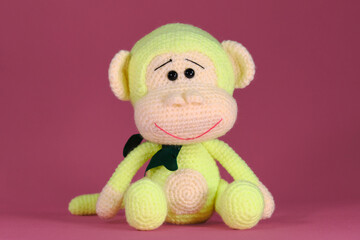 Soft DIY toy made of natural cotton and wool. Cute little monkey with bow on neck crocheted, handmade art. Amigurumi one green monkey with big ears sits on pink background. Front view.