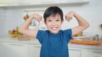 Asian little cute boy in blue shirt demonstrating his muscles or biceps at kitchen room. Boy power...