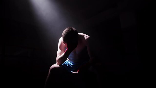 Young caucasian male boxer with wrapped hands sitting in a dark room in a ray of light and looking disappointed. Sportsman is not satisfied with the results of the boxing training