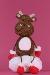 Cute little cow crocheted, handmade art. Amigurumi one brown bull wears red mittens and slippers and sits on white ball of thread on pink background. Soft DIY toy made of natural cotton and wool.