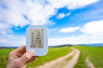 air quality monitor in hand against the background of a green summer field shows the parameters of...