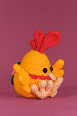 Amigurumi doll chicken with multicolored tail on pink background. Soft DIY toy made of cotton and natural wool. Orange cockerel with bright red comb on its head crocheted, handmade art.