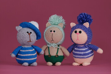 Amigurumi kittens dolls on a pink background stand together. A soft DIY toy made of cotton. Three funny different cats in clothes and funny hats, handmade art.