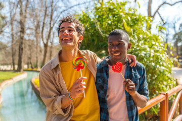 Multi ethnic gay male couple eating a lollipop, lgbt concept, having fun and smiling in a park