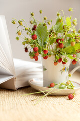 ripe red berries of wild strawberries on  twigs  collected in bunch in tea cup next to open book