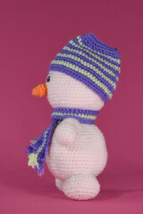 Soft DIY toy made of natural cotton and wool. Crocheted, handmade art. Amigurumi one small white snowman with orange nose and striped hat and scarf on pink background. Side view.