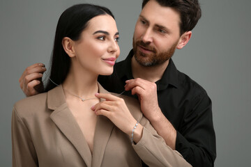 Man putting elegant necklace on beautiful woman against dark grey background