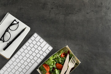 Container of tasty food, keyboard, glasses, cutlery and notebook on grey table, flat lay with space for text. Business lunch