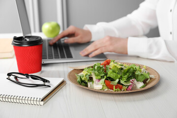Obraz na płótnie Canvas Office employee working with laptop at white wooden table, focus on vegetable salad and paper cup of coffee. Business lunch