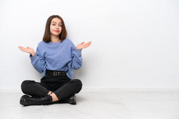 Young Ukrainian woman sitting on the floor isolated on white background having doubts while raising hands