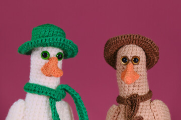 Amigurumi duck dolls on pink background. Soft DIY toy made of cotton. Two brothers goose white with orange beak wears green hat and scarf and brown, crocheted from natural wool, handmade art.