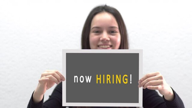 Conceptual message "Now Hiring" on canvas frame label hold by beautiful girl smiling at camera