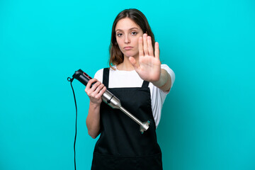 Young caucasian cooker woman using hand blender isolated on blue background making stop gesture