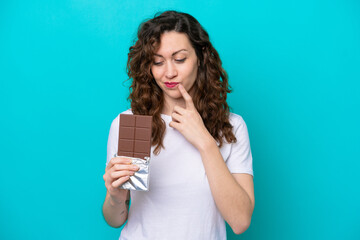Young caucasian woman isolated on blue background taking a chocolate tablet and having doubts