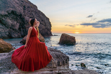 Woman in a red flying dress on the ocean or on the sea beach against the backdrop of the sunset sky.