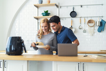 Middle age couple smiling and hugging each other cooking toast bread healthy with recipe by Air Fryer machine,using laptop computer while ecommerce shopping online in grocery store in kitchen at home.