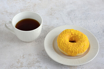 A white plate with a yellow donut on it next to a cup of coffee on marble background