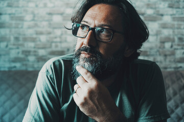 Close up portrait of thoughtful man touching his chin and looking up with eyewear at home on the sofa. Adult lonely man thinking for a solution. Mature people and family economy difficulties thoughts