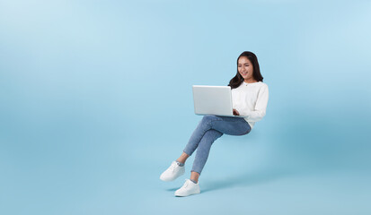 Young smiling Asian girl student floating in mid-air with using laptop isolated on blue studio copy...